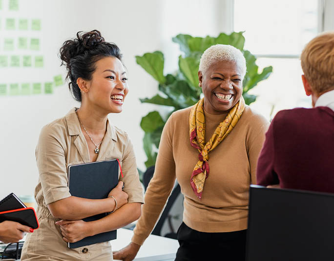 Colleagues standing in a small group discussing something while laughing  Two of the women are holding notebooks 