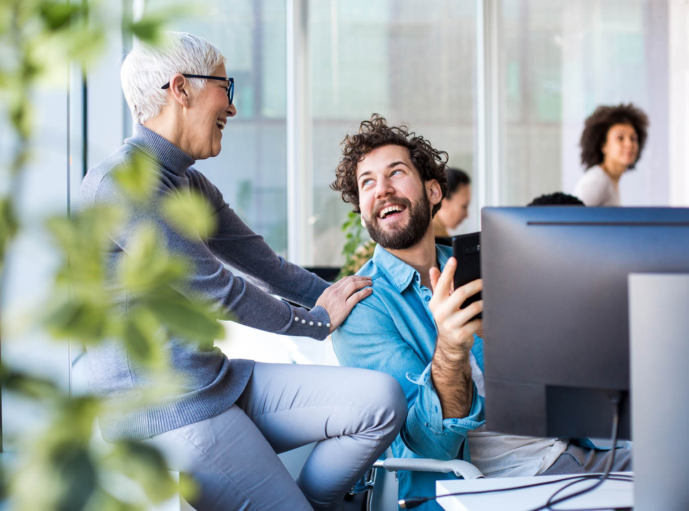 Two office workers taking a minute break in their office  Younger colleague is talking to his mentor and they are positive and smiling  He is sitting at his desk, holding his mobile phone 